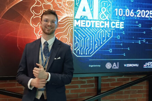 Smiling man in a navy suit and glasses standing in front of AI & MedTech CEE 2025 conference screen showing event date and tech-themed background