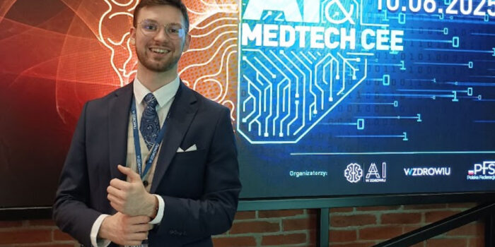 Smiling man in a navy suit and glasses standing in front of AI & MedTech CEE 2025 conference screen showing event date and tech-themed background