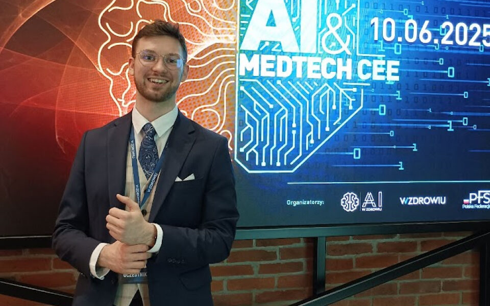 Smiling man in a navy suit and glasses standing in front of AI & MedTech CEE 2025 conference screen showing event date and tech-themed background